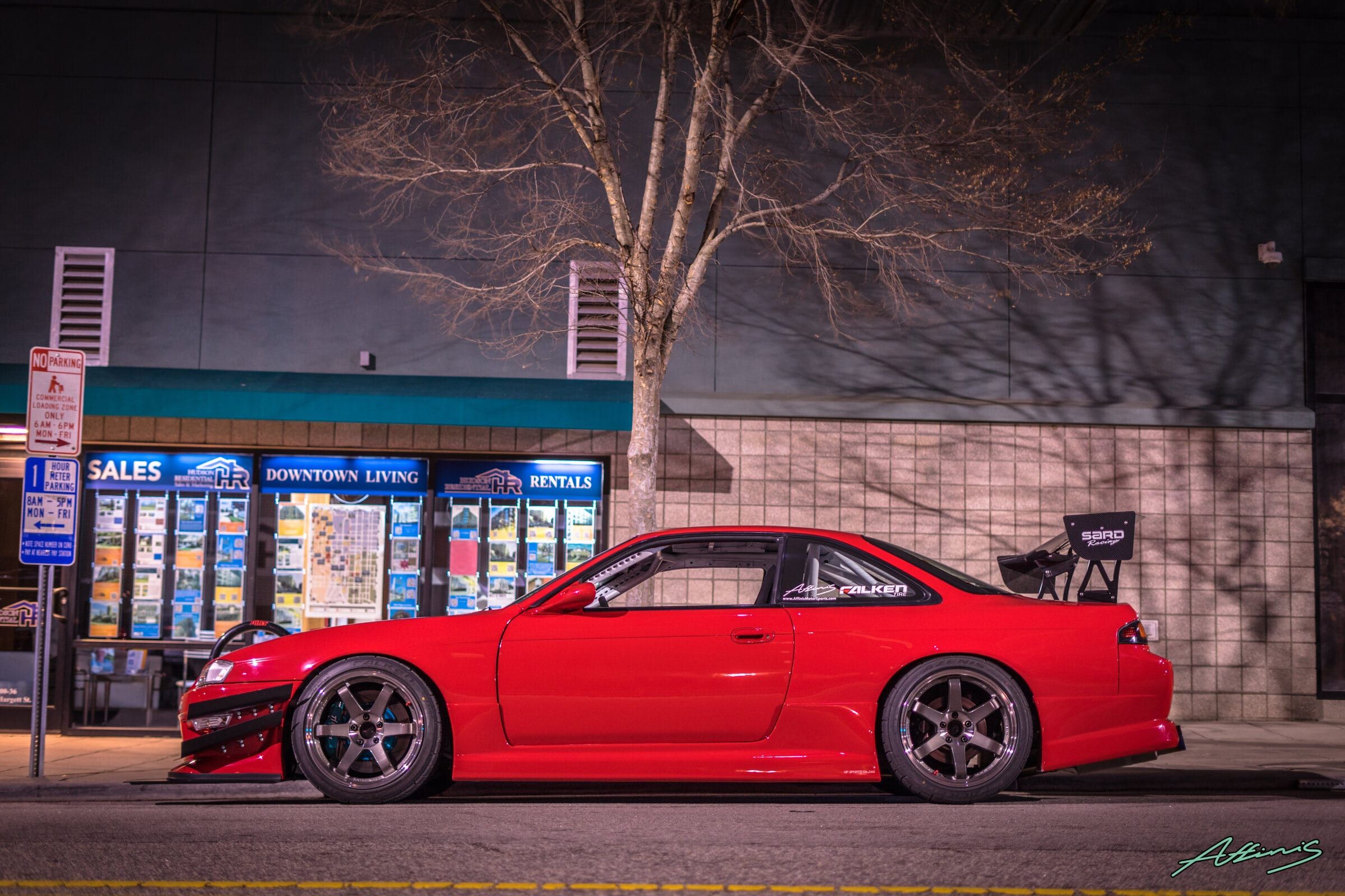 A red Nissan Silvia S14 with a SARD wing parked on a downtown street at night